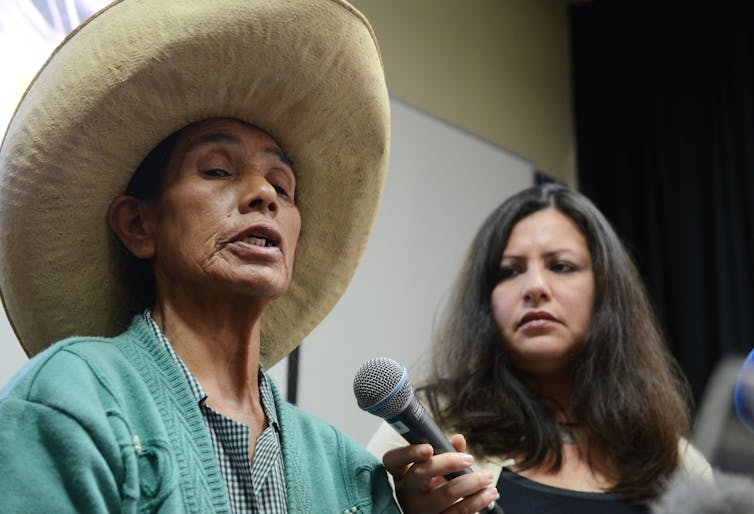 Older woman in green shirt and straw hat speaks into a microphone held by a younger woman