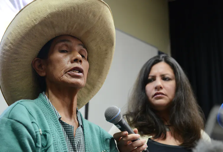 Older woman in green shirt and straw hat speaks into a microphone held by a younger woman