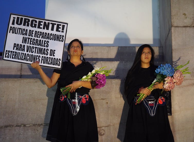 Women in black holding flower bouquets and signs stand solemnly in front of a wall