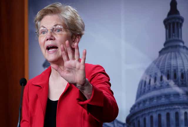 Sen. Elizabeth Warren holds up her left hand as she speaks during a news conference, with an image of the Capitol dome in the background