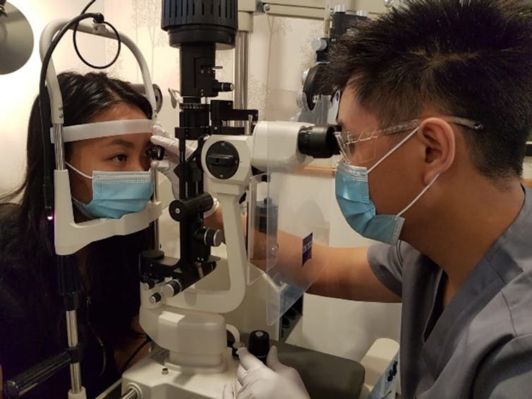 Ophthalmologist wearing face mask, glasses and gloves, performing eye exam with patient.