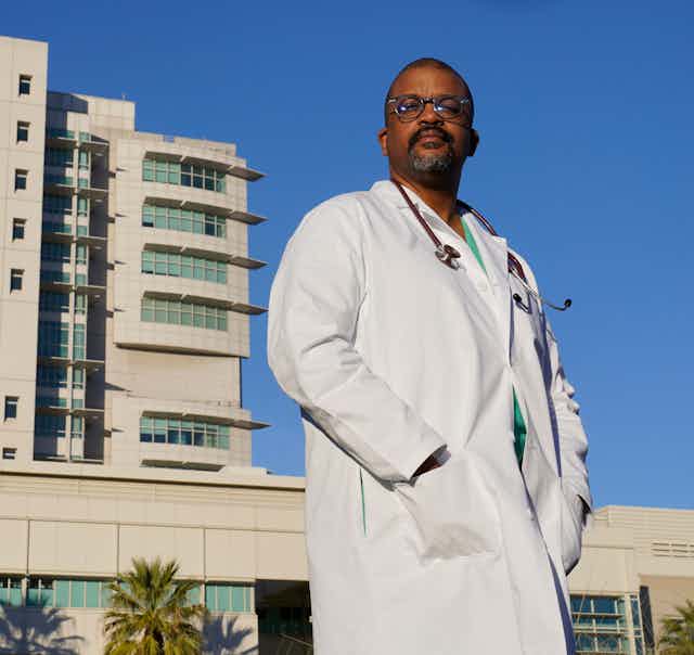 A doctor in his white coat stands in front of his hospital