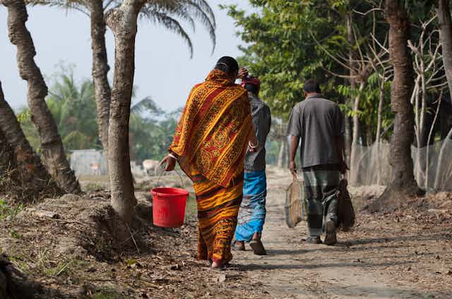 Three people walk on a dirt path by palm trees carrying buckets