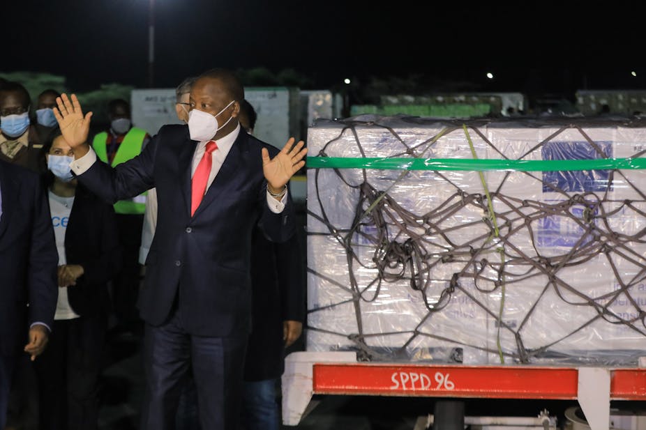 At night, a man in a face mask and a smart suit with a red tie holds his hands up in a pleased gesture, cargo on a trolley behind him.