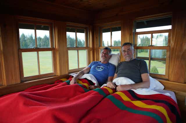 Two men lie in a bed with a red cover, and views out the many windows shows trees, grass and nature