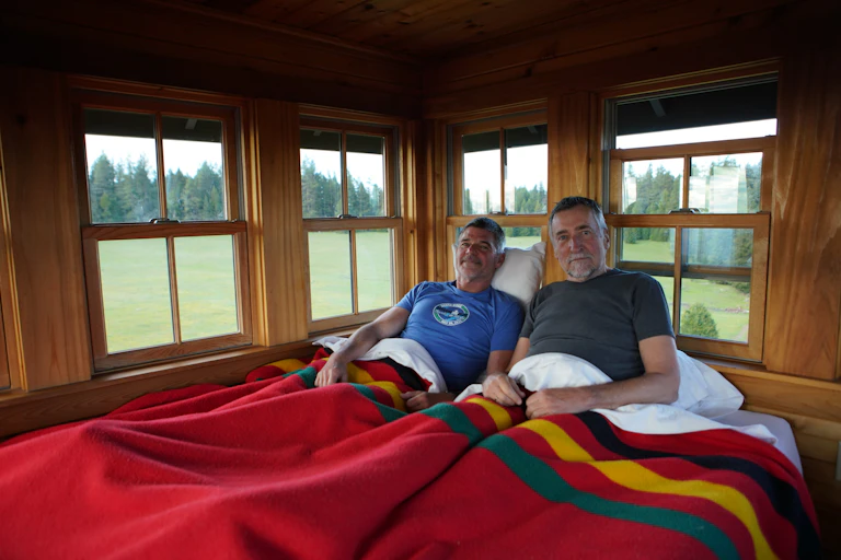 Two men lie in a bed with a red cover, and views out the many windows shows trees, grass and nature