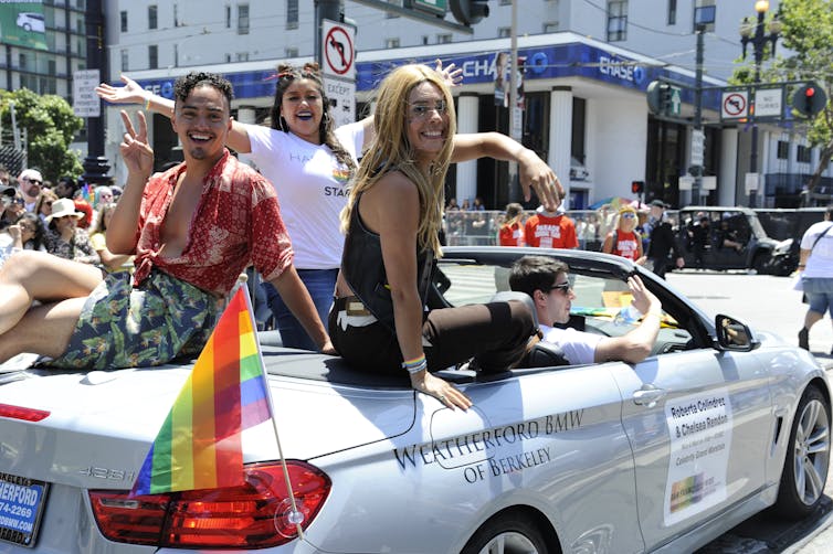 Three smiling LGBTQ Latinos ride along a city street in an open-top convertible with a rainbow flag