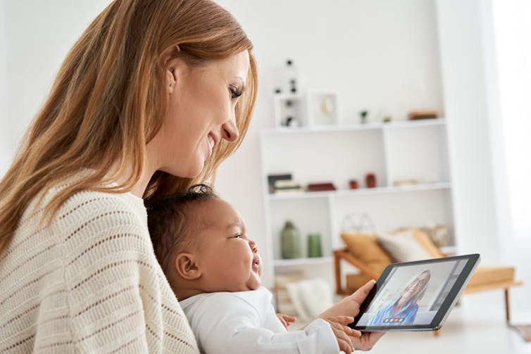 A woman and her baby look at an older woman on a tablet.