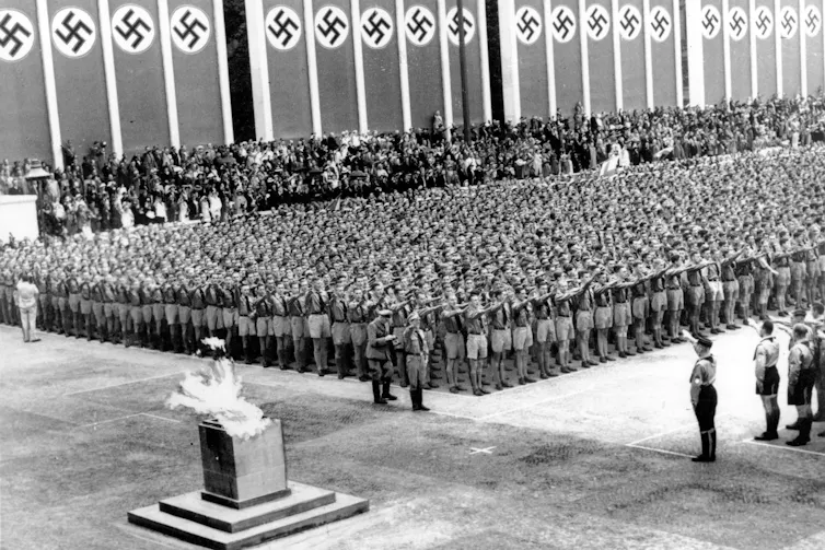 Thousands of soldiers give the Nazi salute while the Olympic flame burns in the foreground.
