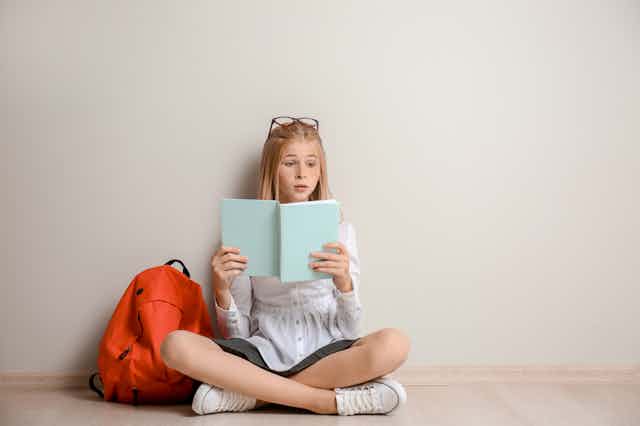Girl sitting on floor reading a blue book next to orange backpack
