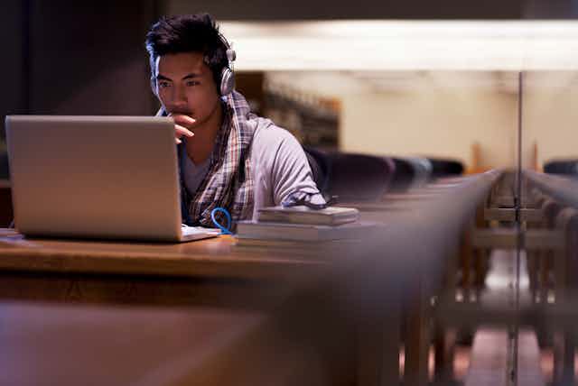 A man wearing headphones looks at his laptop in an empty library.