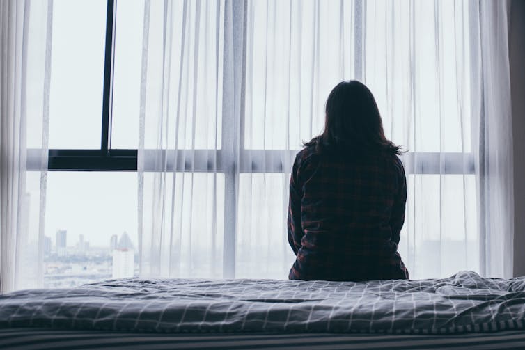 A woman sits on a bed looking out the window.