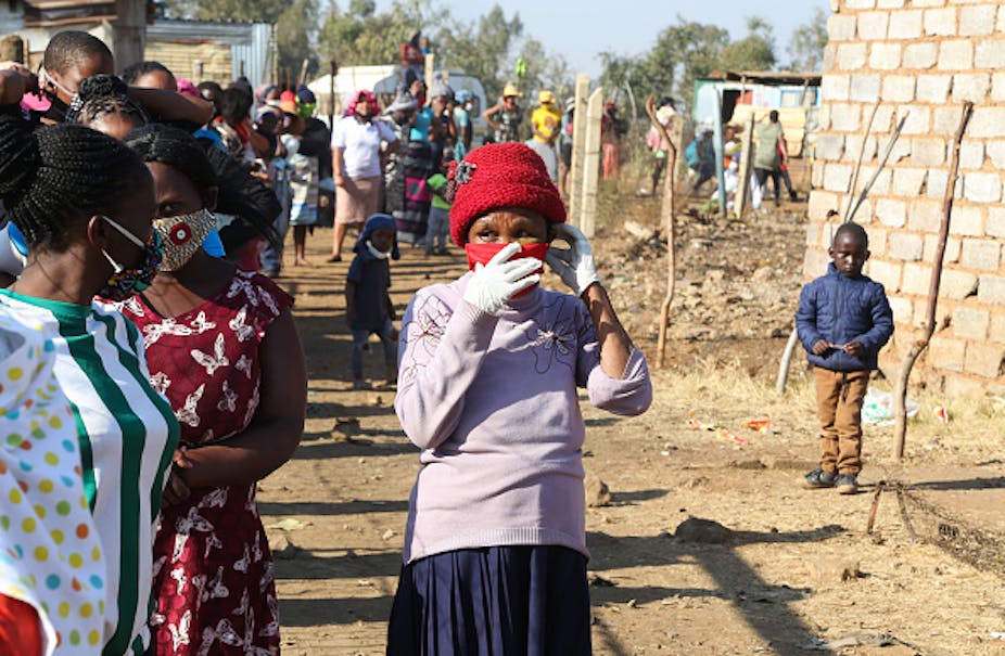 People wearing face masks stand in a line, with shacks in the background