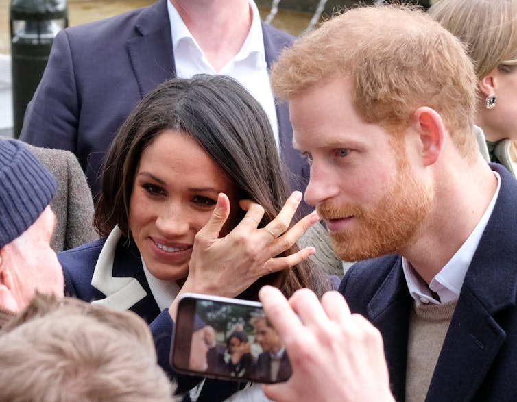 Harry and Meghan are photographed on a smartphone while speaking to a crowd