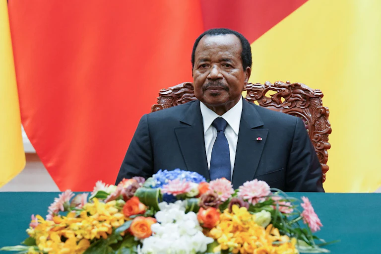 An old man wearing a dark suit, white shirt and dark tie sits behind a table adorned with flowers