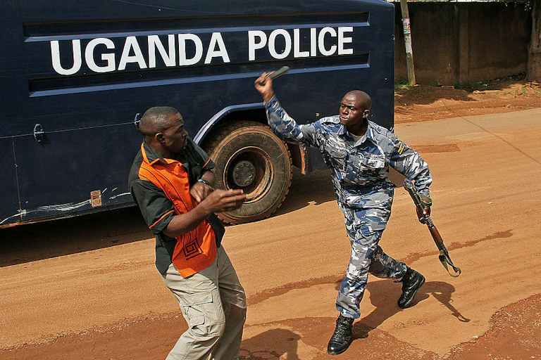 A policeman, rifle in left hand, aims a club at the face of an unarmed journalists with his right.