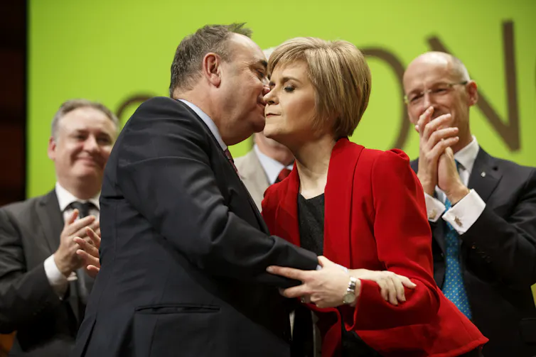 Alex Salmond embraces Nicola Sturgeon surrounded by clapping colleagues.