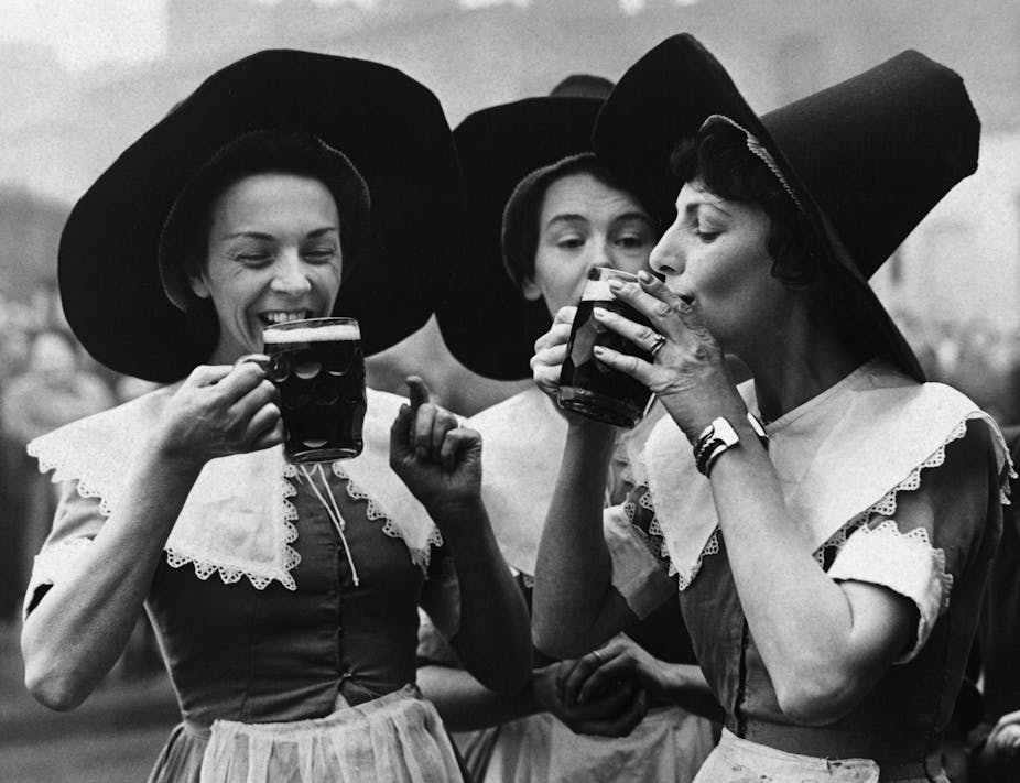 Three women wearing tall hats smile and sip from mugs of beer.