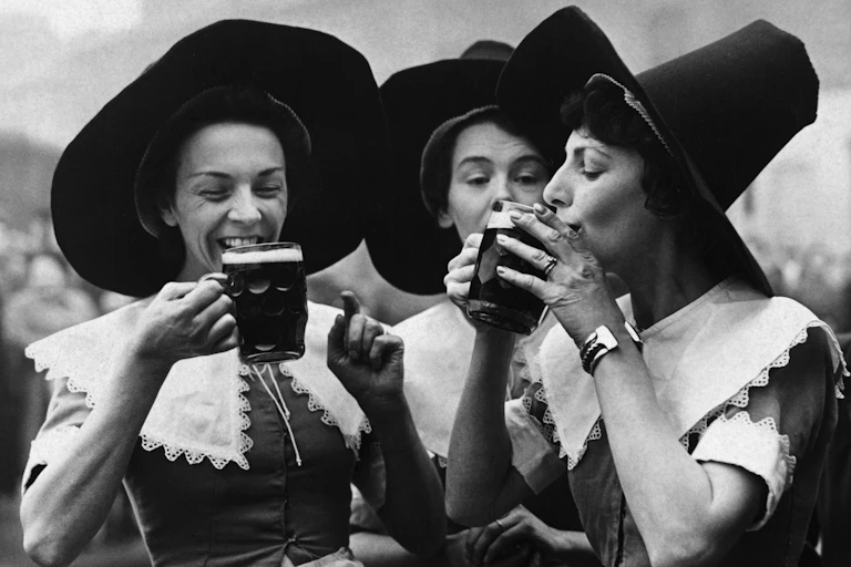 Three women wearing tall hats smile and sip from mugs of beer.