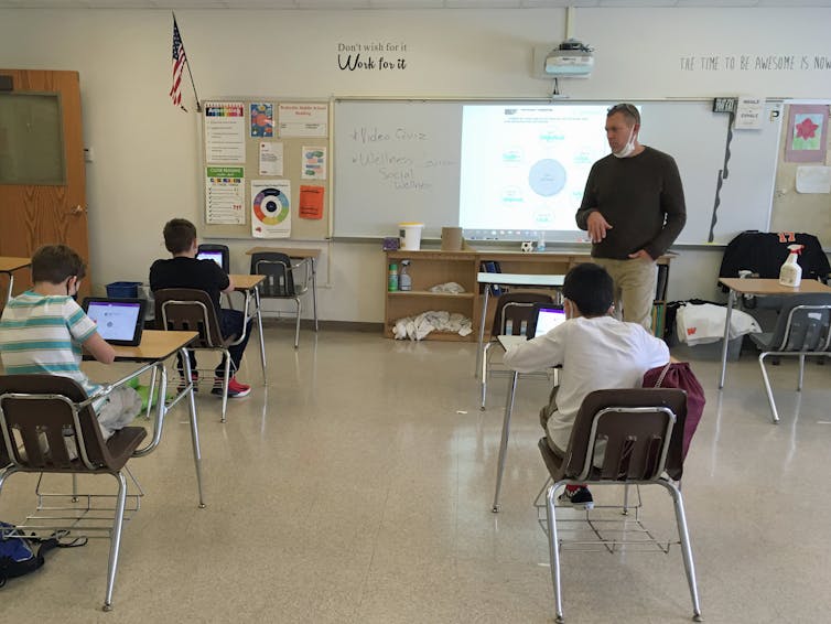 A teacher wearing a mask conducts his class while his students sit using iPads.