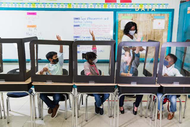 A teacher wearing a mask calls on her students who are sitting in desks with plastic shields.