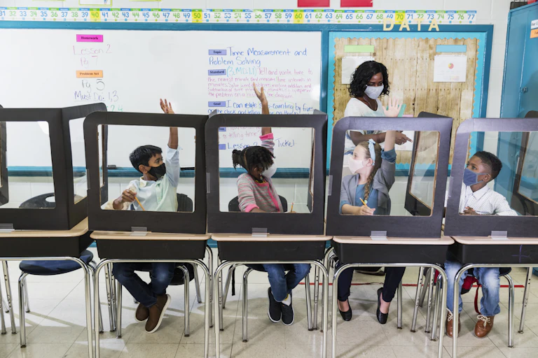 A teacher wearing a mask calls on her students who are sitting in desks with plastic shields.