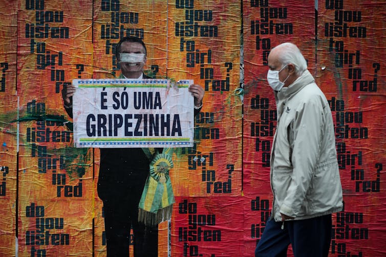 A man in a mask walks next to an image of Brazilian President Jair Bolsonaro holding a sign with his comment about COVID-19 at the beginning of the pandemic, 'It's just a little flu,'