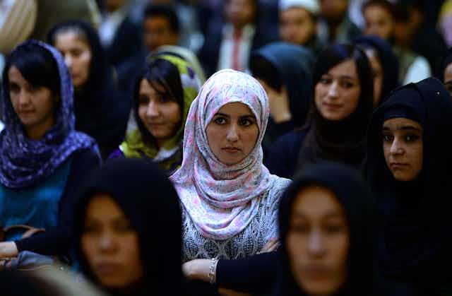 Women, most in hijab, stand in a crowd and listen attentively. 