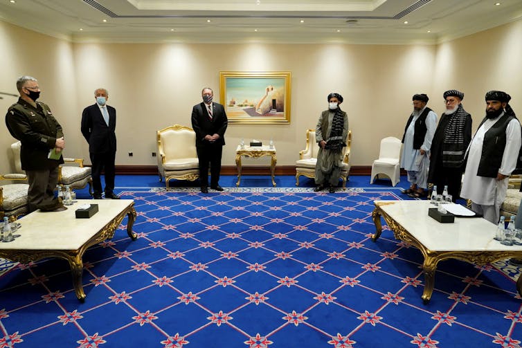Men, some in suits and other in traditional Pashtun clothing, stand in a hotel conference room at a distance from each other, wearing face masks