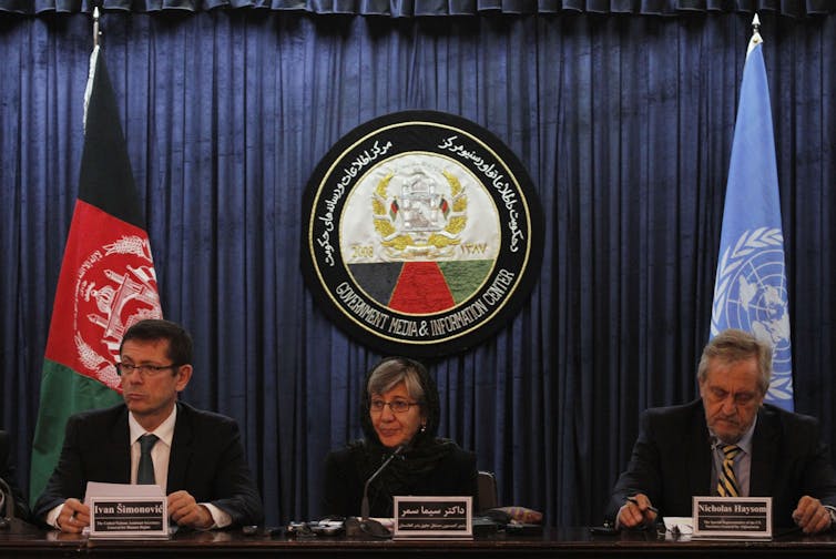 Woman in hijab sits at a table with microphones between two men in sits, with international flags behind them