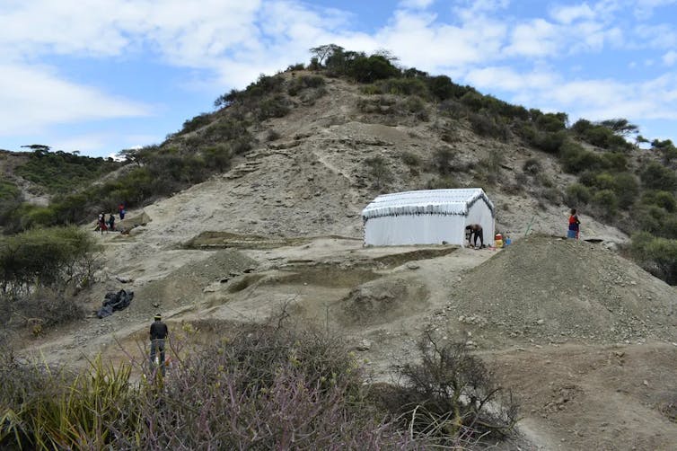 Archaeologists working next to a tent on a hillside.