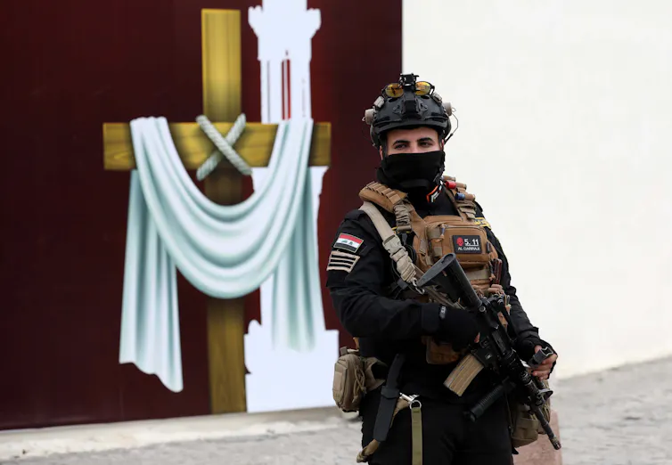 A soldier holding a rifle stands guard over a church in Baghdad. In the background is a cross decorated with a white robe.