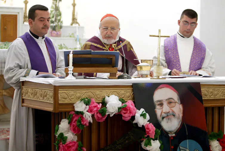 Three priests celebrate funeral mass in church in Baghdad. In front of altar is photograph of murdered archbishop Paulos Faraj Rahho.