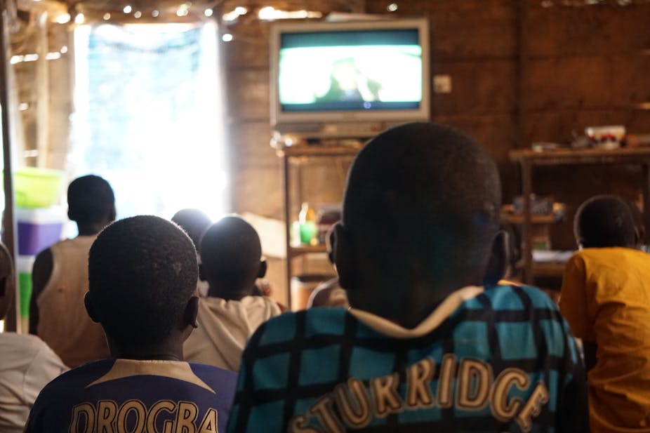An informal structure with lights hanging above the door; children sit in rows watching a television set, their backs to the camera.