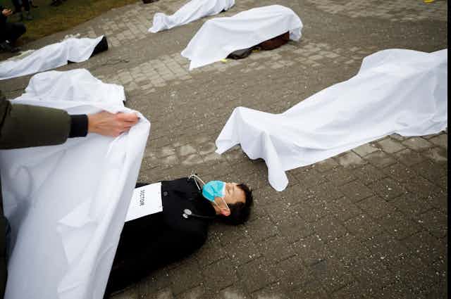 A person lies on the ground as someone covers them with a white sheet with others covered by white sheets behind him.