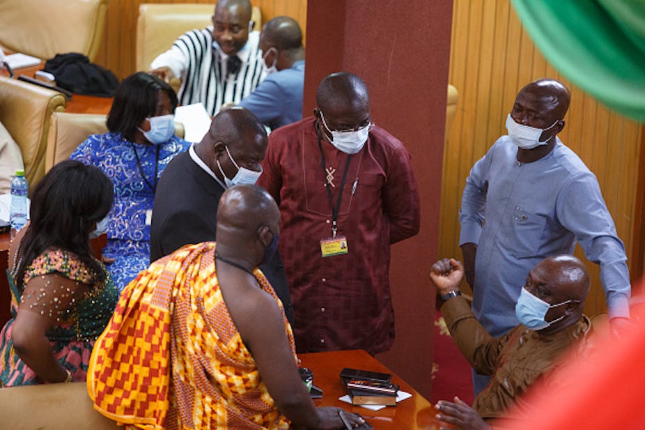 In the foreground, a circle of seven people - five of them men - in smart clothing in parliament in Ghana; they wear face masks but are clearly deep in debate, a man who is seated commanding the attention of the others.