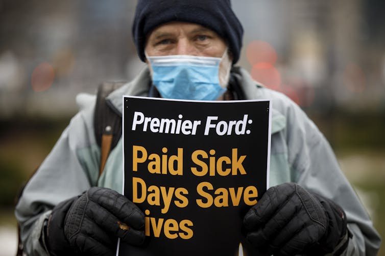 A man wearing a mask holds up a sign that reads Doug Ford: Paid Sick Days Save Lives