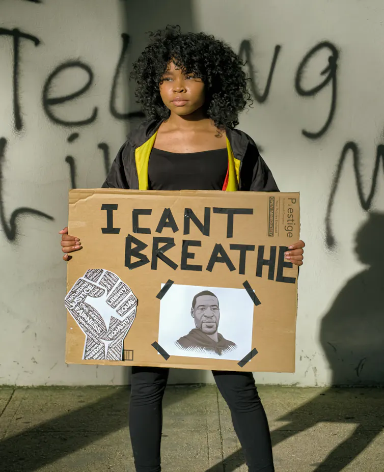 A girl holds a cardboard sign with a picture of George Floyd and text reading I CANT BREATHE