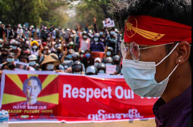 A man in a mask and protest bandana with, in the background, a crowd of protesters on the streets of Yangon, Myanmar.