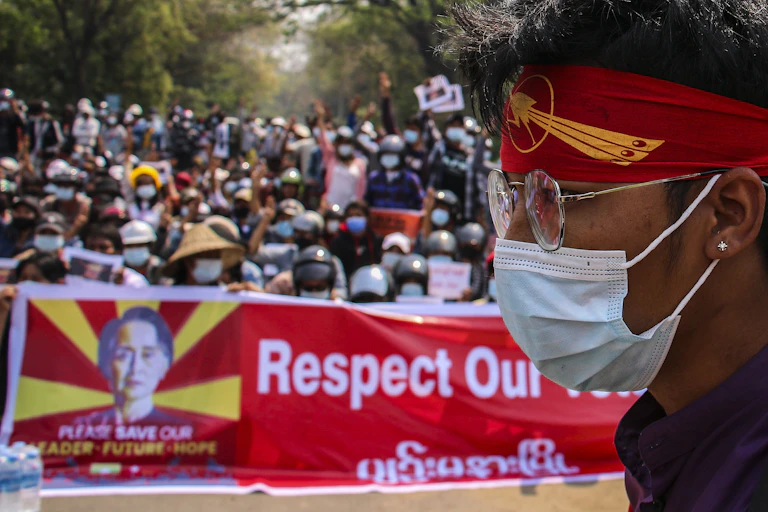 A man in a mask and protest bandana with, in the background, a crowd of protesters on the streets of Yangon, Myanmar.