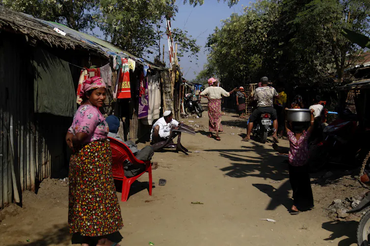 Rohingya people sit and walk by a temporary building.
