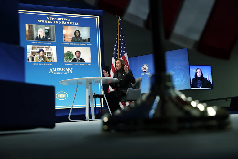 Kamala Harris, wearing black, sits by a big video screen, at a table with hands raised while speaking to a virtual forum on 'supporting women and families'