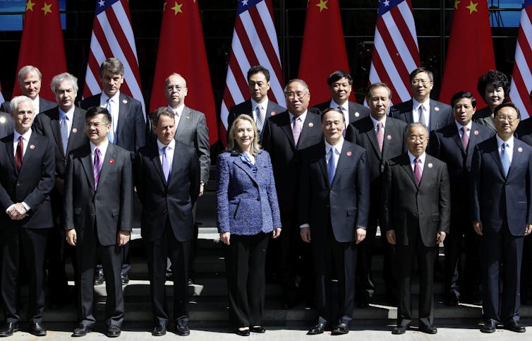 Clinton, in a blue blazer, stands at the center of a large group of Chinese and American diplomats posing for a picture, all wearing black suits