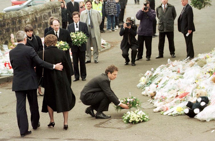 Tony Blair placing a wreath in front of a large pile of flowers with other mourners and photographers.