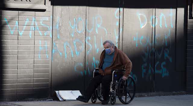 A man wearing a face mask wheels his wheelchair past a spray-painted wall that reads Wash Your Hands Don't Touch Your Face.