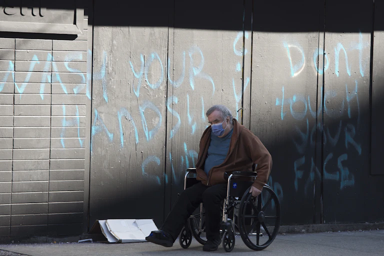 A man wearing a face mask wheels his wheelchair past a spray-painted wall that reads Wash Your Hands Don't Touch Your Face.