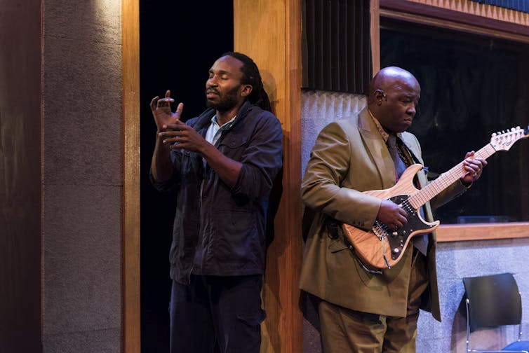 Two black men: one mimics playing a trumpet, one plays guitar.