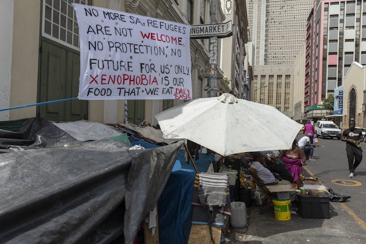 Sign written in black and red with text that reads: 'No more SA, refugees are not welcome. No protection, no future for us only xenophobia is our food that we eat daily basis', next to parasol covering refugee shelter