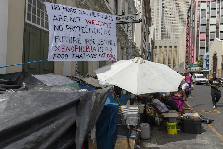 Sign written in black and red with text that reads: 'No more SA, refugees are not welcome. No protection, no future for us only xenophobia is our food that we eat daily basis', next to parasol covering refugee shelter