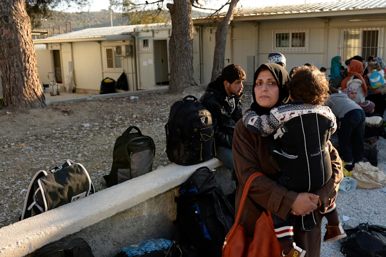 Muslim woman looking into camera while carrying baby with rucksacks and other refugees in the background